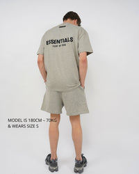 Rear view of a man wearing the Fear of God Essentials Dark Oatmeal Summer set with a loose-fitting short-sleeve top and matching shorts, paired with black sneakers, against a plain white background.