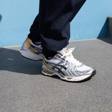 Person wearing ASICS Gel-Kayano 14 White Pure Gold sneakers standing on pavement against a blue wall, showcasing the shoe’s white and gold design with black accents, perfect for exclusive sneaker enthusiasts in Europe. The real deal.