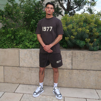 Man wearing dark gray Fear of God Essentials 1977 Iron Summer Set with 1977 printed on both shirt and shorts, paired with white sneakers against a greenery backdrop.