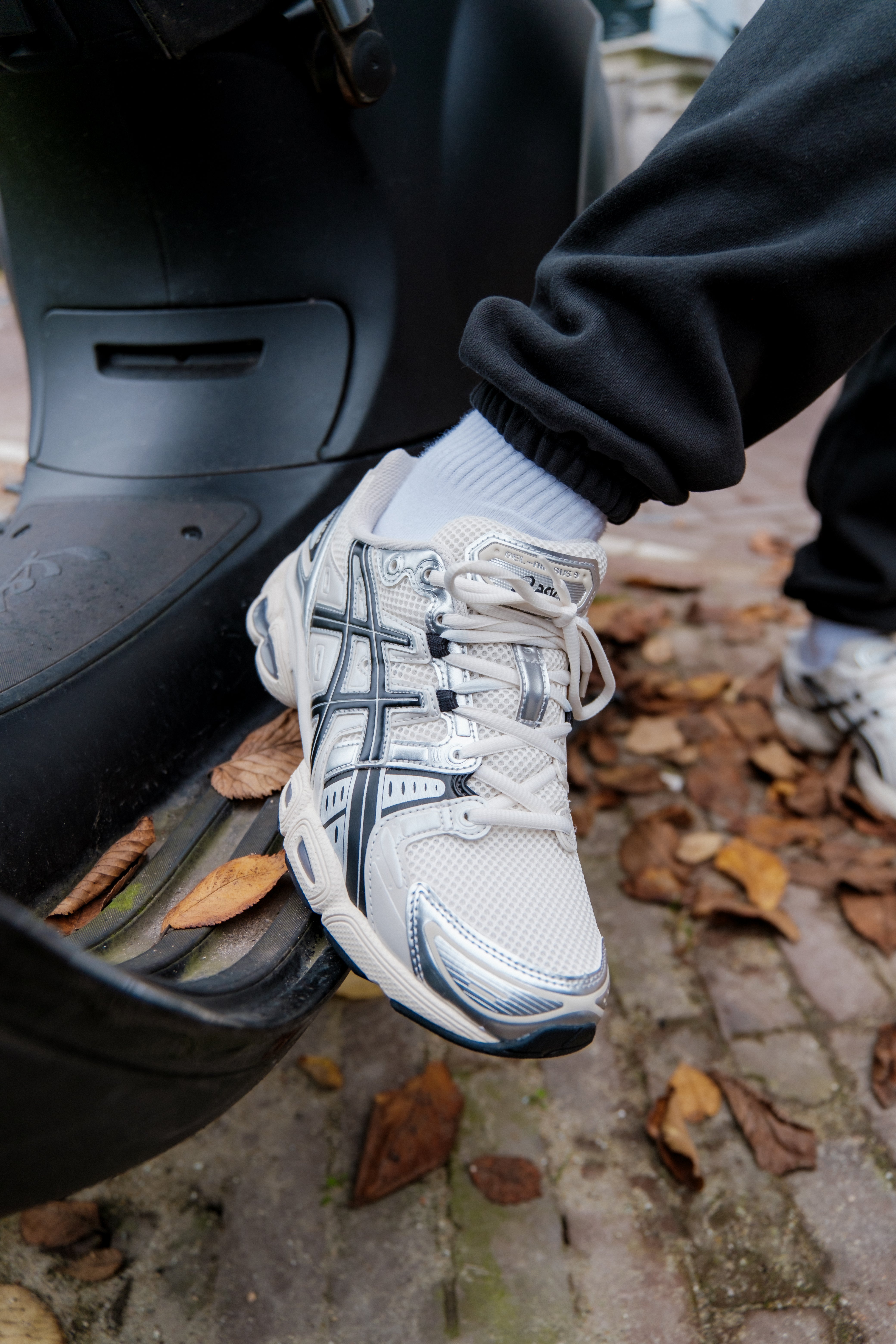 Close-up of a person wearing Asics Gel-Nimbus 9 Cream Black sneakers with black joggers and white socks, stepping on a scooter footboard surrounded by autumn leaves, showcasing the classic sneaker design. The real deal.