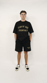 A man wearing the Fear of God Essentials FW24 Black Yellow Summer Set consisting of a black oversized t-shirt with yellow lettering and matching black shorts, paired with black and white sneakers, standing against a plain white background.