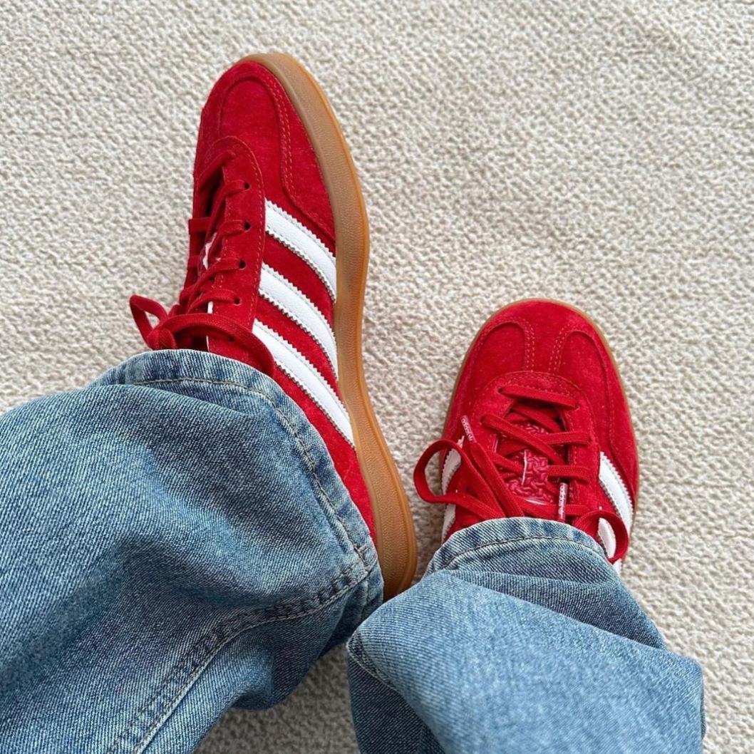 Person wearing Adidas Gazelle Scarlet Cloud White sneakers with red suede upper and white stripes, paired with blue jeans on a light carpet, showcasing exclusive trainers Europe.