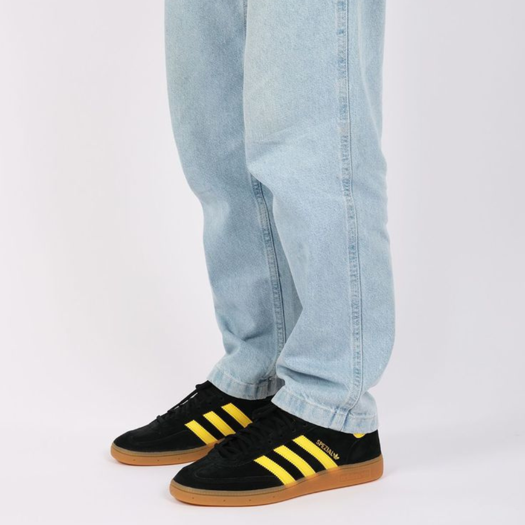 Person wearing Adidas Handball Spezial Black Yellow sneakers with black suede upper and yellow stripes, paired with light blue jeans, standing on a white background.