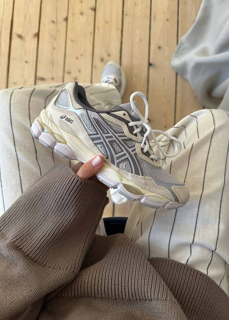 Person wearing a beige and white striped outfit holding an ASICS Gel-NYC Oatmeal sneaker with white laces and grey detailing on a wooden floor, showcasing exclusive European sneakers.