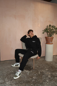 Man sitting on a chair wearing Fear of God Essentials FW24 Black White Lange Broek with black hoodie and white sneakers, in a minimalist room with a potted plant and textured floor, showcasing exclusive sneakers in whole Europe. The real deal.