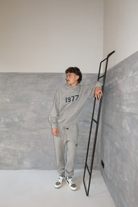 Man wearing Fear of God Essentials 1977 Dark Oatmeal Lange Broek sweatpants and matching hoodie, posing casually against a modern concrete wall while leaning on a black ladder, showcasing exclusive clothing style.