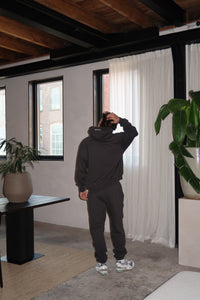 Man wearing Fear of God Essentials 1977 Iron Black Lange Broek standing indoors with hand on head, modern casual outfit paired with sneakers, urban minimalist setting.