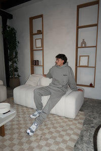 Man sitting on white couch wearing Fear of God Essentials Dark Oatmeal Lange Broek gray sweatpants and matching hoodie with white sneakers in a modern living room setting.