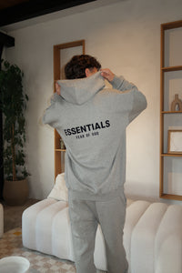 Man wearing a grey hoodie with "ESSENTIALS FEAR OF GOD" text on the back, paired with Fear of God Essentials Dark Oatmeal Lange Broek pants in a modern, minimalist living room setting, showcasing exclusive sneakers and clothing style, the real deal.