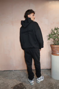 Man wearing a black Fear of God Essentials Stretch Limo Hoodie with matching pants and white sneakers, standing indoors near a plant against a textured beige wall.