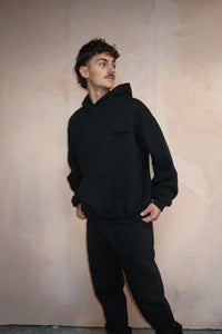 Man wearing a black Fear of God Essentials Stretch Limo Full set with a relaxed fit hoodie and matching joggers, posing against a neutral wall, showcasing exclusive European clothing.