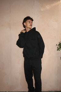 A man wearing black Fear of God Essentials Stretch Limo Lange Broek posing against a beige textured wall with minimal decor in a casual, stylish setting.