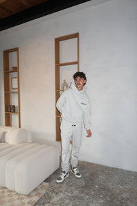 Man wearing a Fear of God Essentials Light Oatmeal Hoodie paired with matching sweatpants and sneakers, standing in a minimalist room with neutral tones, showcasing casual streetwear style.