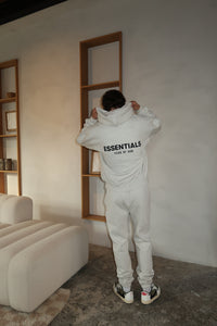 Person wearing Fear of God Essentials Light Oatmeal Lange Broek standing indoors facing a wall, dressed in matching light oatmeal pants and hoodie with black lettering on the back.