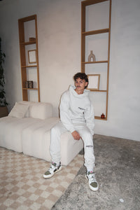 Young man wearing Fear of God Essentials Light Oatmeal Lange Broek seated on a modern cream sofa in a minimalist living room with neutral tones and wooden shelving.