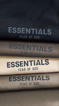 Close-up of folded clothing featuring printed text "ESSENTIALS FEAR OF GOD" in various muted colors, highlighting the Fear of God Essentials Off Black Korte Broek product.