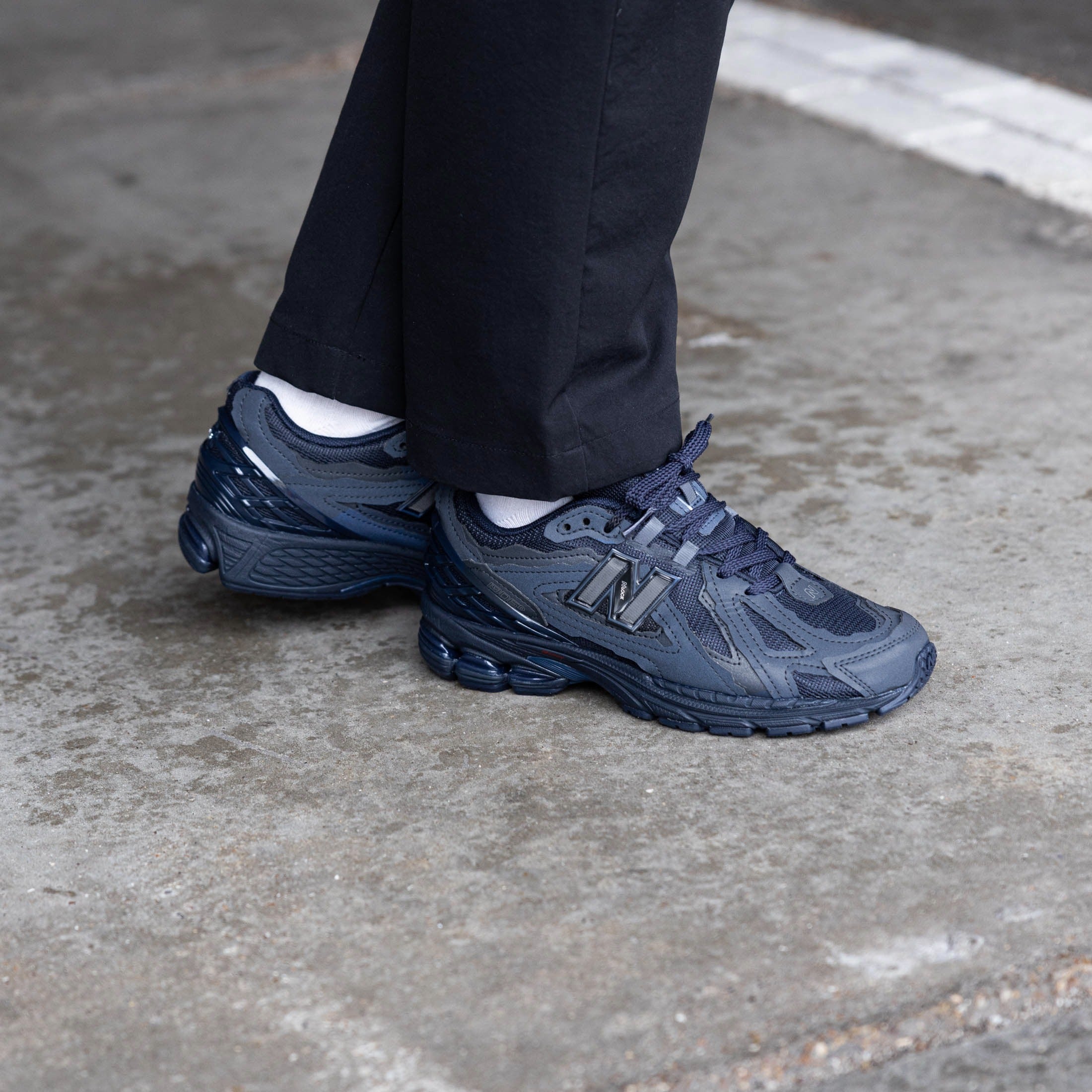 Side view of a person wearing dark navy blue New Balance 1906D Protection Pack Eclipse sneakers paired with black pants and white socks, standing on a concrete surface, showcasing exclusive European sneakers – The real deal.