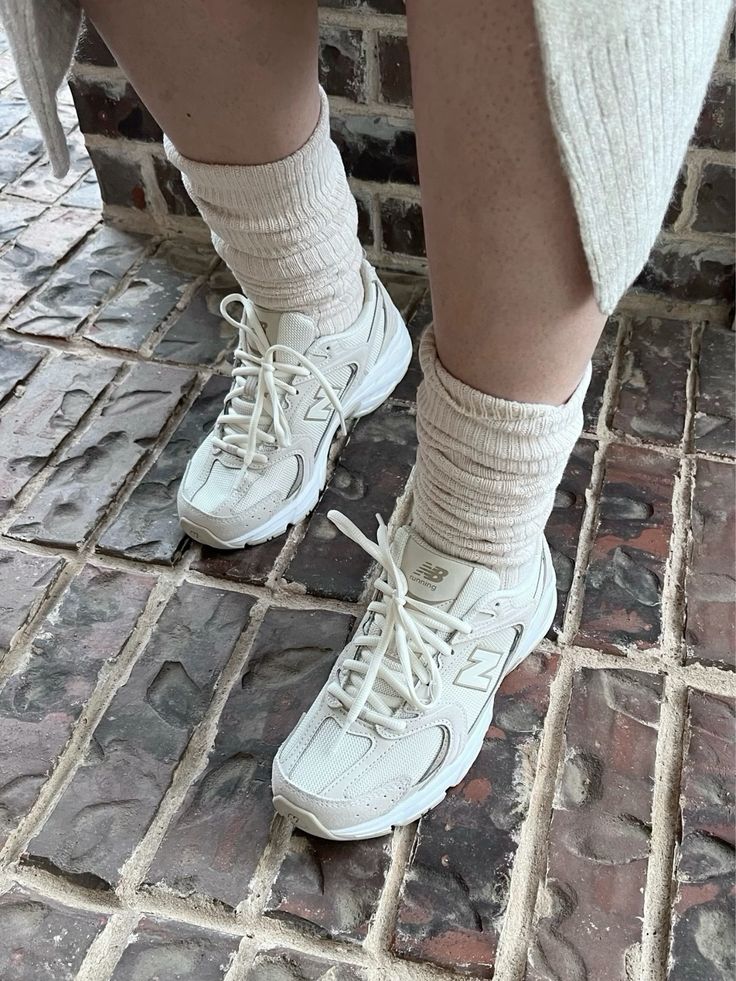 A person wearing cream-colored socks and New Balance 530 Sea Salt Moonbeam sneakers, standing on a textured stone floor, showcasing exclusive European footwear.