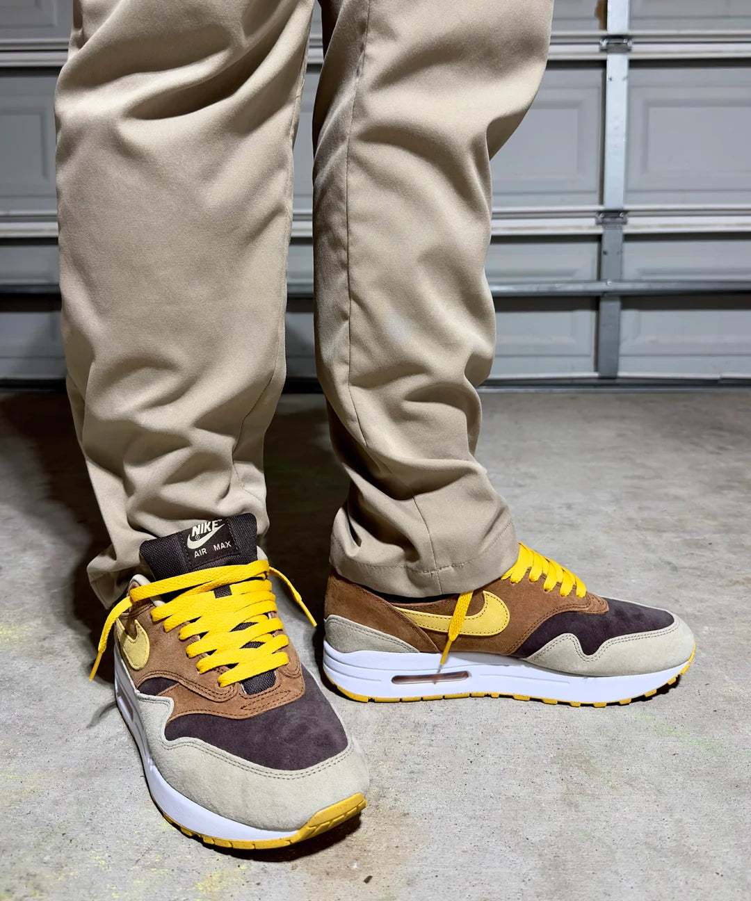 Close-up of feet wearing Nike Air Max 1 PRM Duck Pecan sneakers with yellow laces, showcasing brown and beige tones on a concrete floor, emphasizing exclusive style in Europe The real deal
