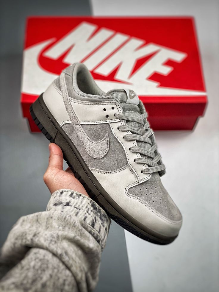 Hand holding a Nike Dunk Low Ironstone sneaker in shades of grey and white with a red Nike shoebox in the background, showcasing exclusive European sneakers – The real deal.