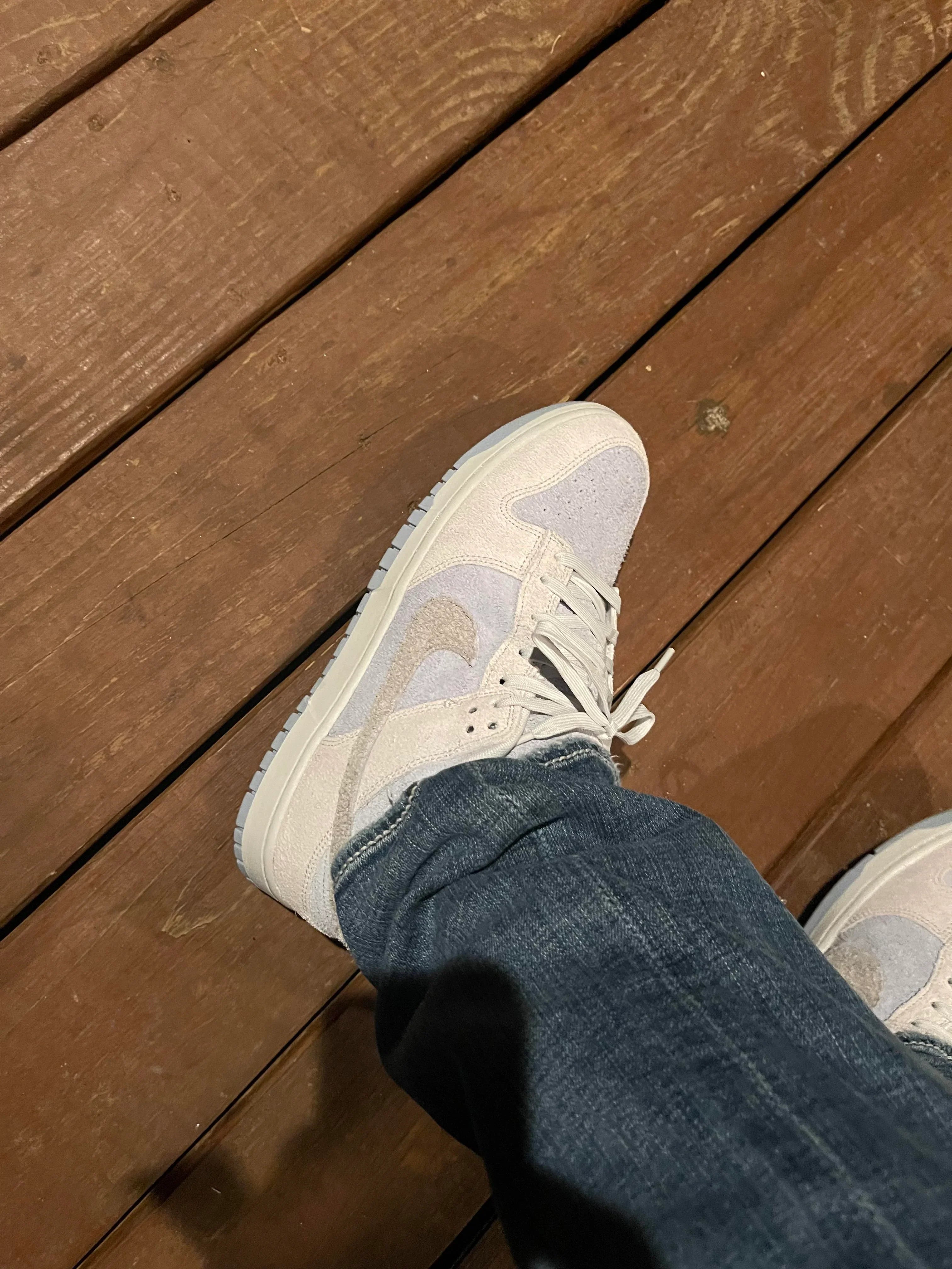 Close-up of a person wearing Nike Dunk Low Light Armory Blue Photon Dust sneakers with light blue and cream hues, paired with blue denim jeans on wooden flooring, the real deal.