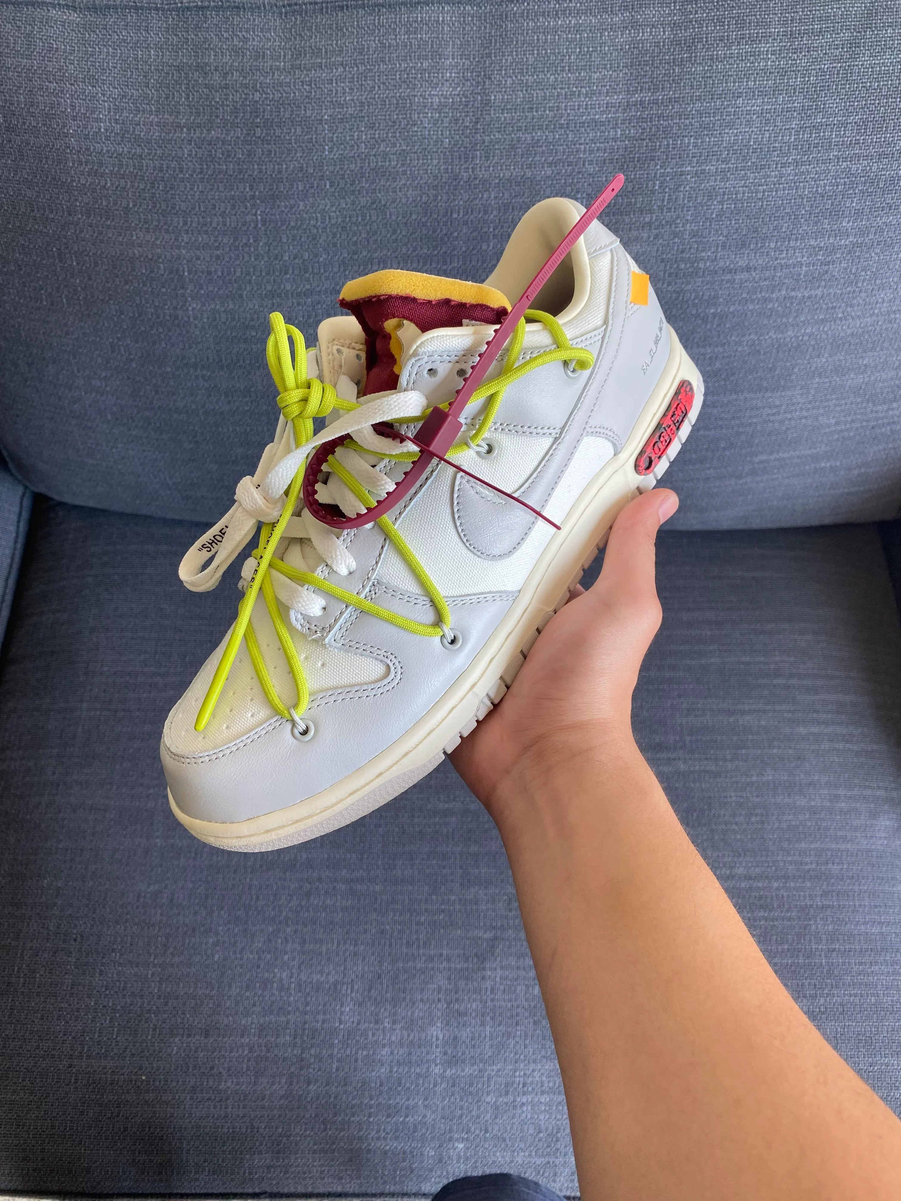 Hand holding a white Nike Dunk Low Off-White Lot 8 sneaker featuring neon yellow laces, exposed foam tongue with maroon accents, and signature Off-White zip tie against a gray fabric background. The real deal.