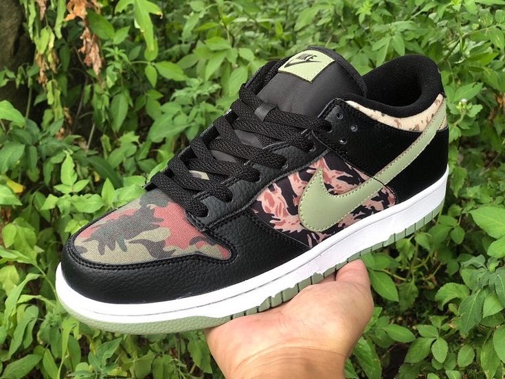 Hand holding a Nike Dunk Low SE Black Multi-Camo (Crazy Camo) sneaker featuring black leather and multicolor camouflage patterns, photographed outdoors among green foliage, the real deal.