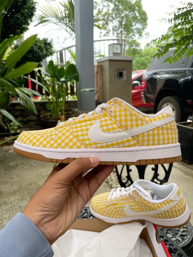 Hand holding a Nike Dunk Low Yellow Gingham sneaker featuring a yellow and white checkered pattern with white swoosh and gum sole, showcased outdoors with another pair in the background, representing exclusive sneakers available in whole Europe. The real deal.