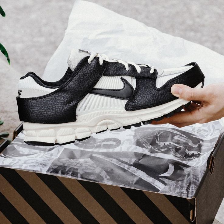 A close-up of a black and white Nike Zoom Vomero 5 Timeless Panda Dunk sneaker held above a shoebox with patterned wrapping paper, highlighting its textured leather and cushioned sole, the real deal exclusive in Europe.