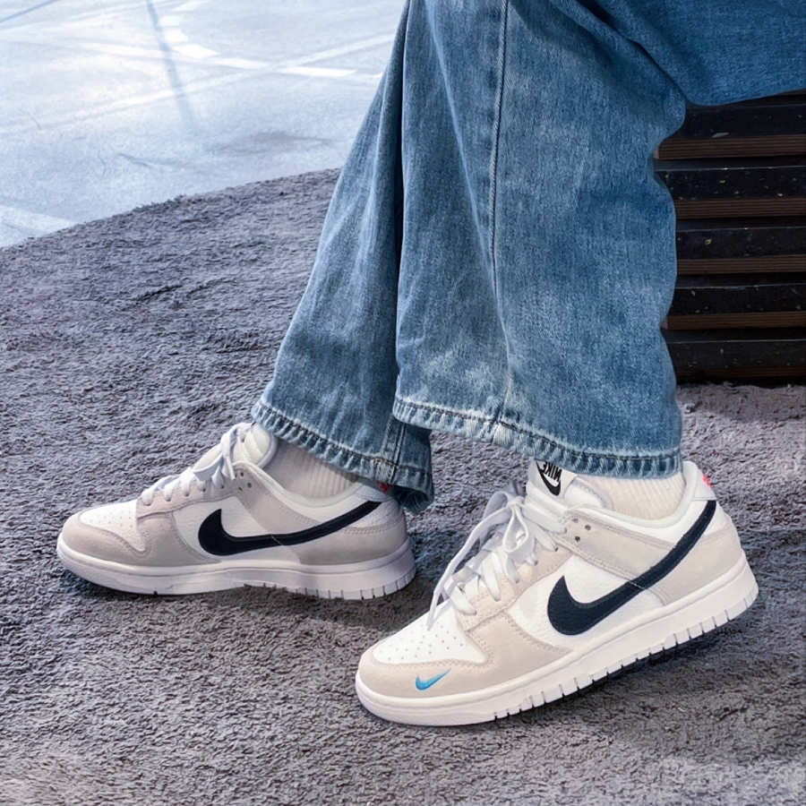 Person wearing Nike Dunk Low White Grey Navy Aqua Mini Swoosh sneakers with white, grey, navy, and aqua accents, paired with blue denim jeans, sitting on a carpeted floor.