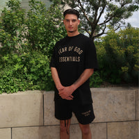 A man outdoors wearing the Fear of God Essentials FW24 Black Yellow Summer Set, featuring a black oversized t-shirt and matching shorts with beige lettering, showcasing casual summer clothing.