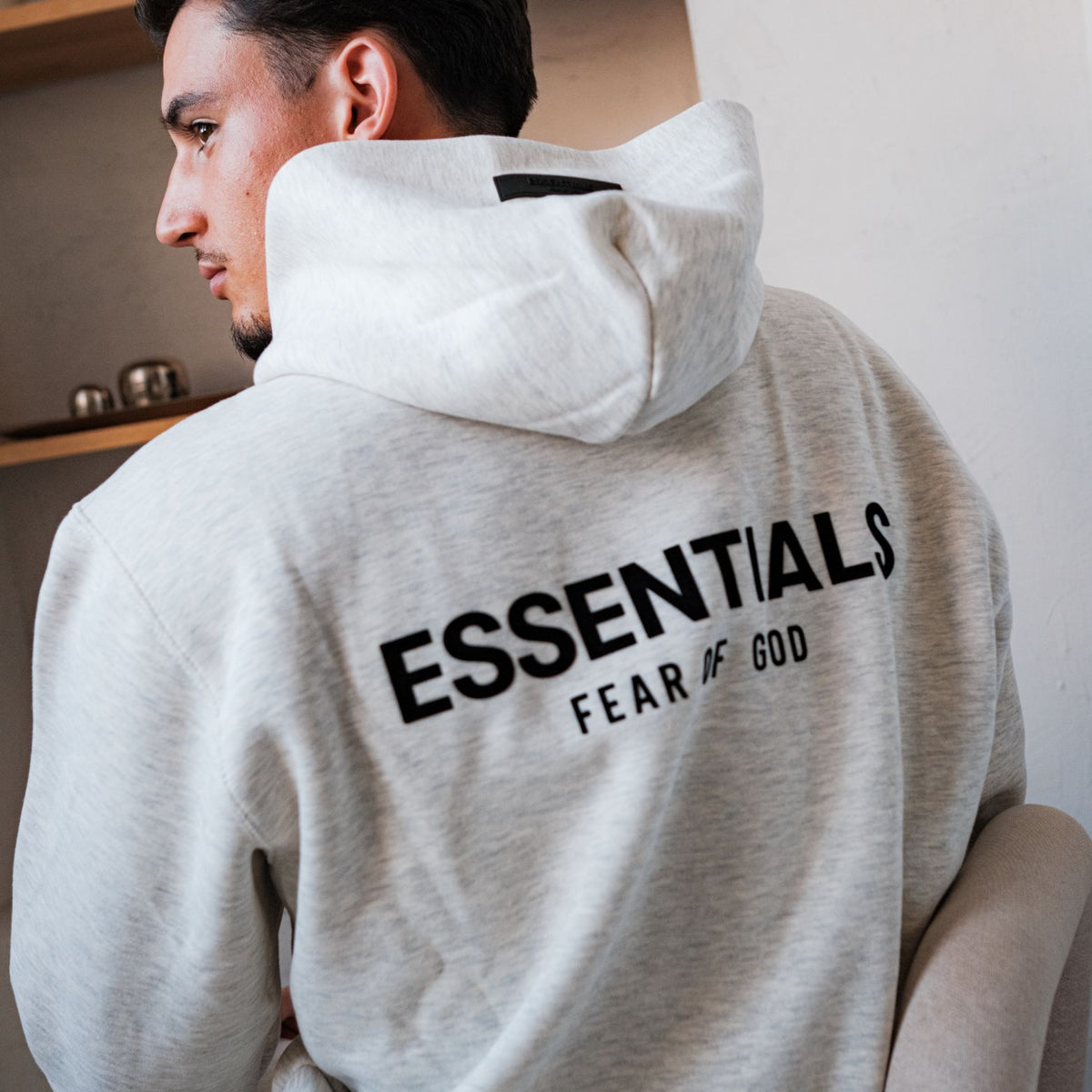 Fear of God Essentials Light Oatmeal Hoodie, achteraanzicht met zwart geborduurd logo en witte capuchon