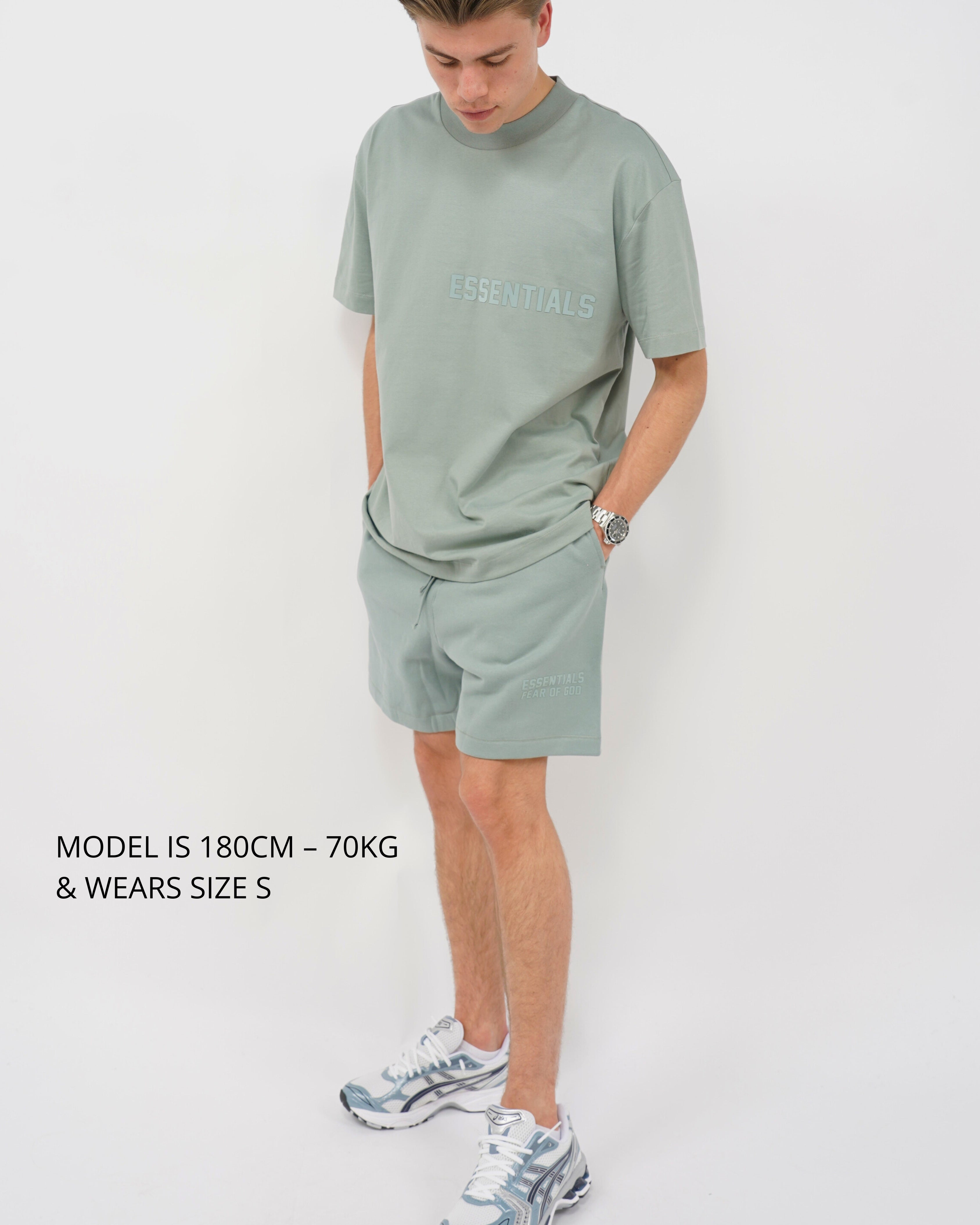 Male model wearing Fear of God Essentials Sycamore Summer set in muted green, styled with casual sneakers, standing against a plain white background in a relaxed pose.