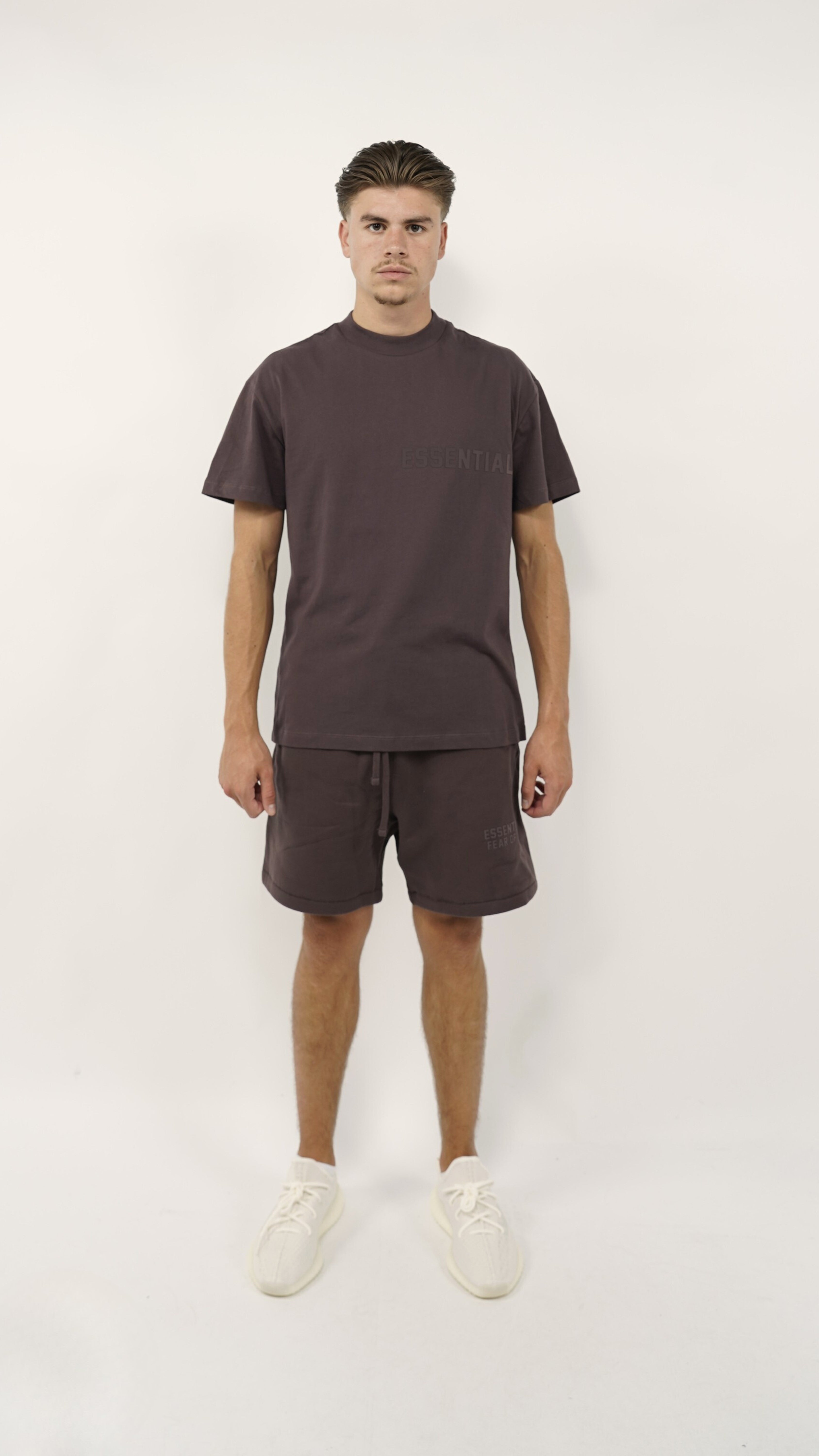 Man wearing Fear of God Essentials Plum Summer set with matching plum-colored shorts and t-shirt paired with white sneakers, showcasing casual summer clothing.