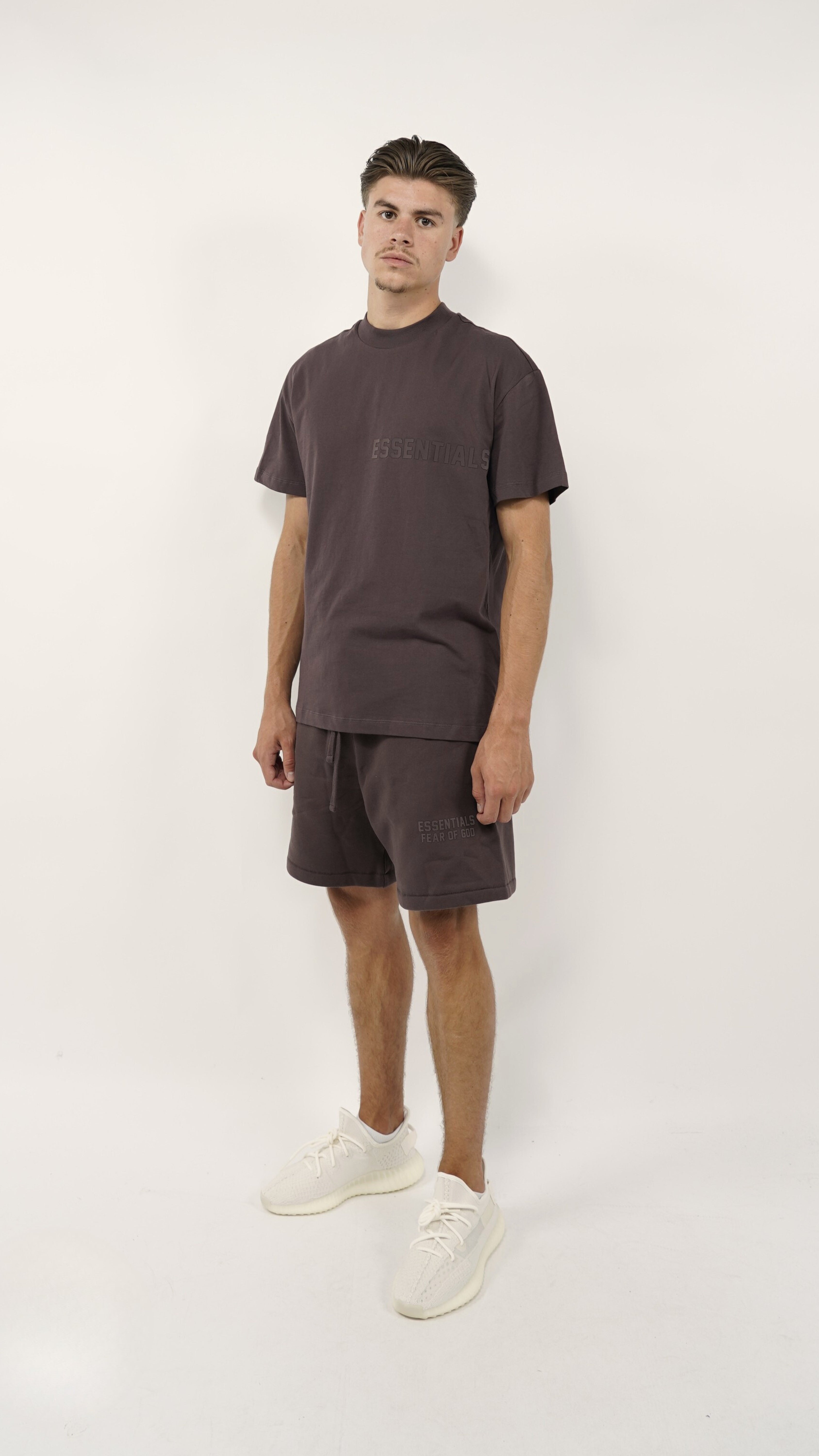 Male model wearing the Fear of God Essentials Plum Summer set with matching plum-colored shirt and shorts paired with white sneakers, standing against a plain background.