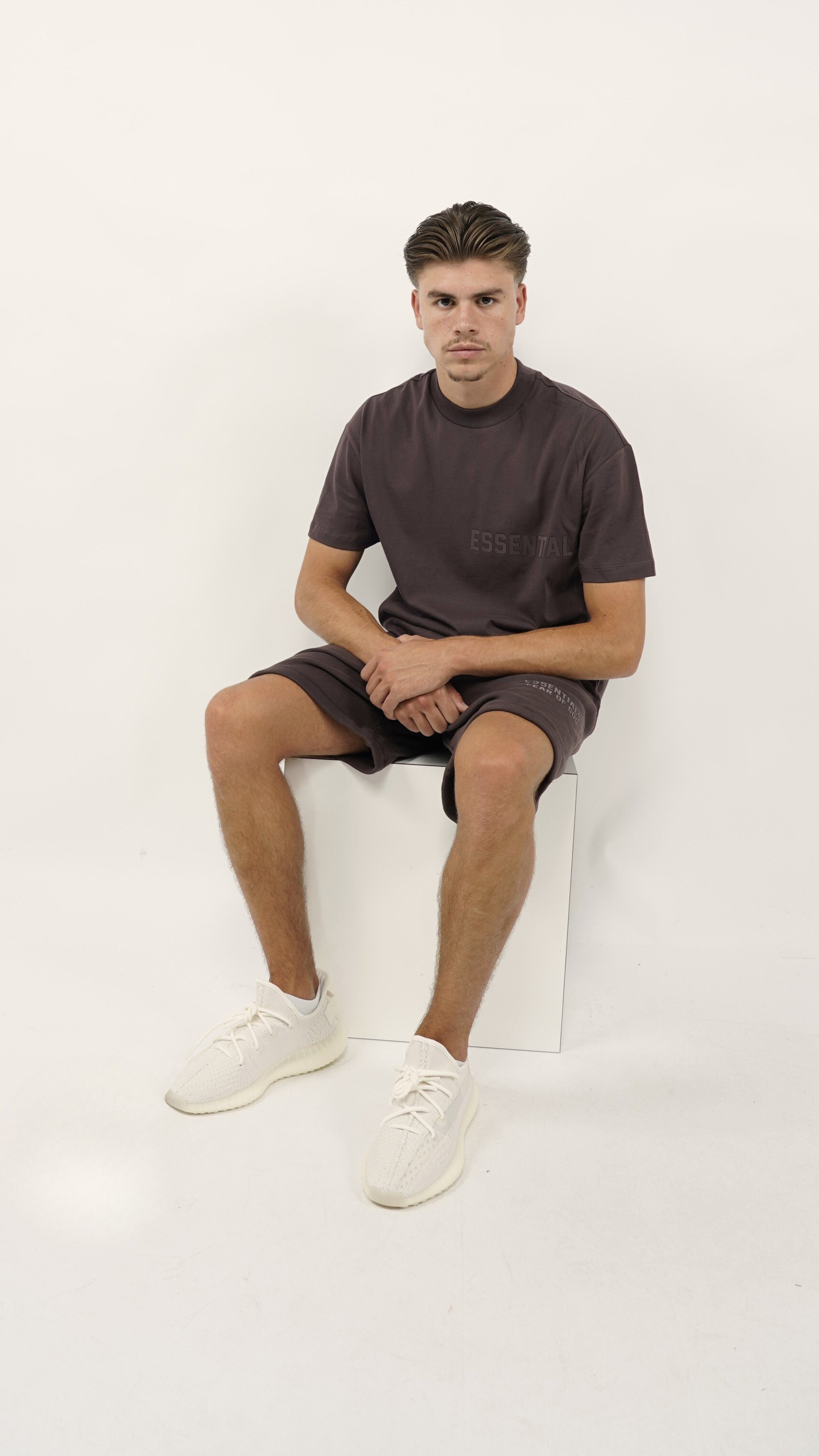 Young man seated wearing the Fear of God Essentials Plum Summer set with matching plum shirt and shorts paired with white sneakers, embodying casual style and comfort.