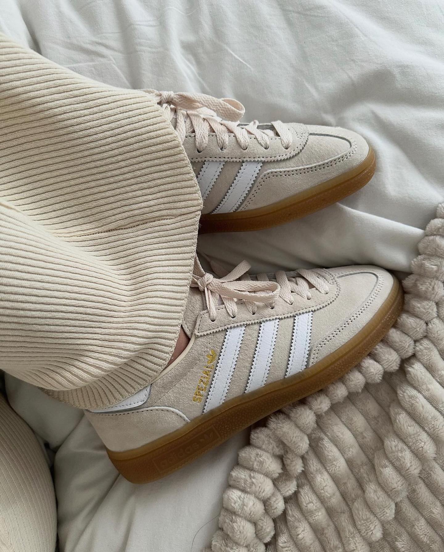 Close-up of a person wearing Adidas Handball Spezial Wonder White sneakers with beige ribbed pants, resting on a cozy cream textured blanket, showcasing a stylish and casual look.