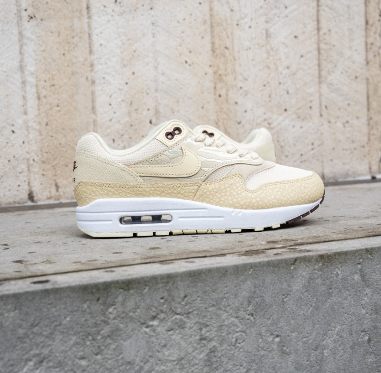 Side view of Nike Air Max 1 '87 Safari Coconut Milk sneakers in beige with textured accents, displayed on a concrete ledge, showcasing exclusive European sneaker style.