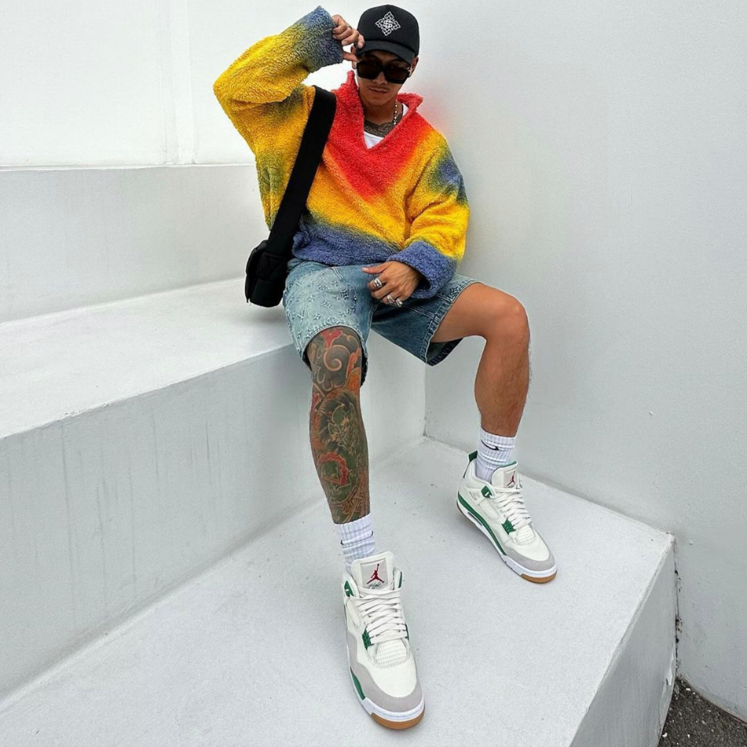 A stylish person wearing colorful knitwear, denim shorts, a black cap, and Nike SB x Air Jordan 4 Retro Pine Green sneakers, showcasing exclusive European sneaker fashion.