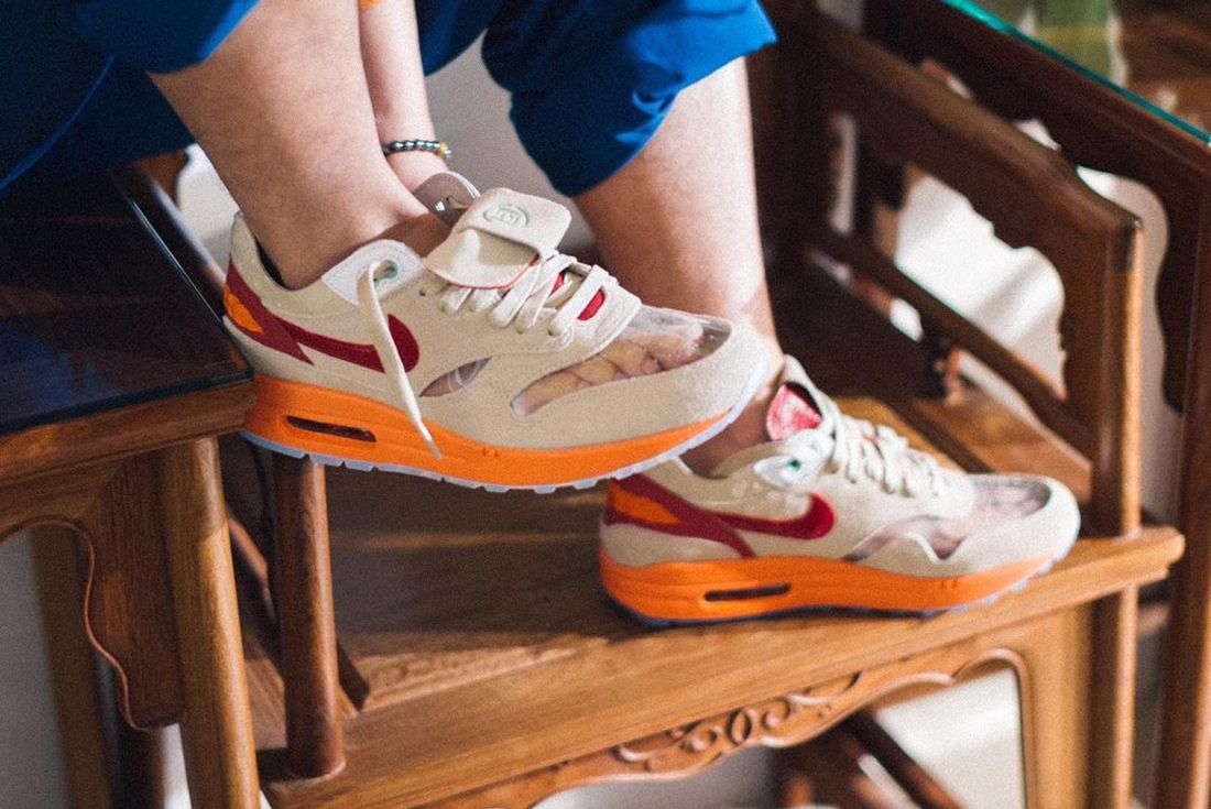 Casual view of a person wearing Nike Air Max 1 Clot Kiss of Death sneakers with beige, orange, and red accents, sitting on a wooden carved chair. The real deal.