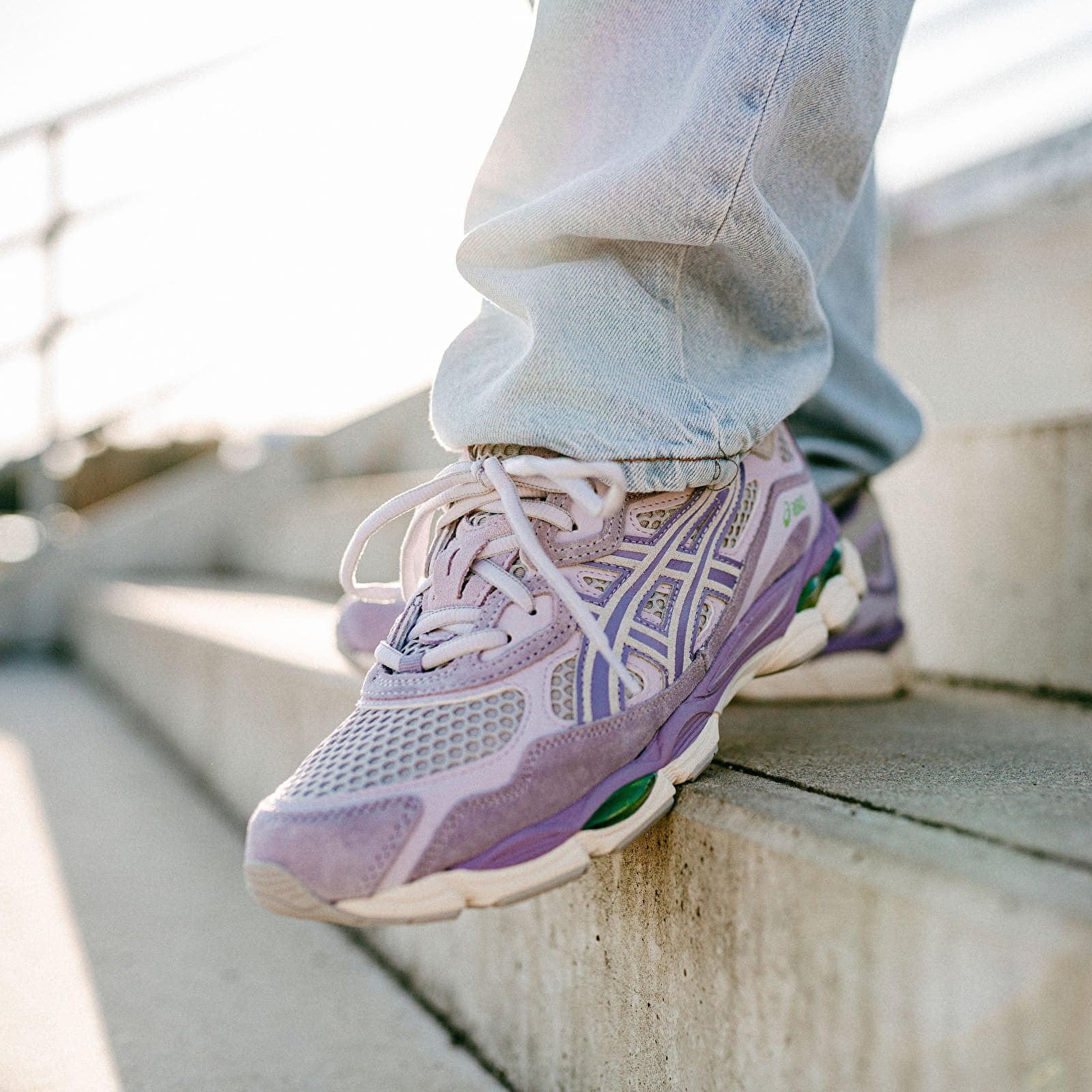 Close-up of feet wearing ASICS Gel-NYC Cement Grey Ash Rock sneakers in soft purple and grey tones, styled with light denim jeans on outdoor concrete steps, showcasing exclusive European trainers – The real deal.