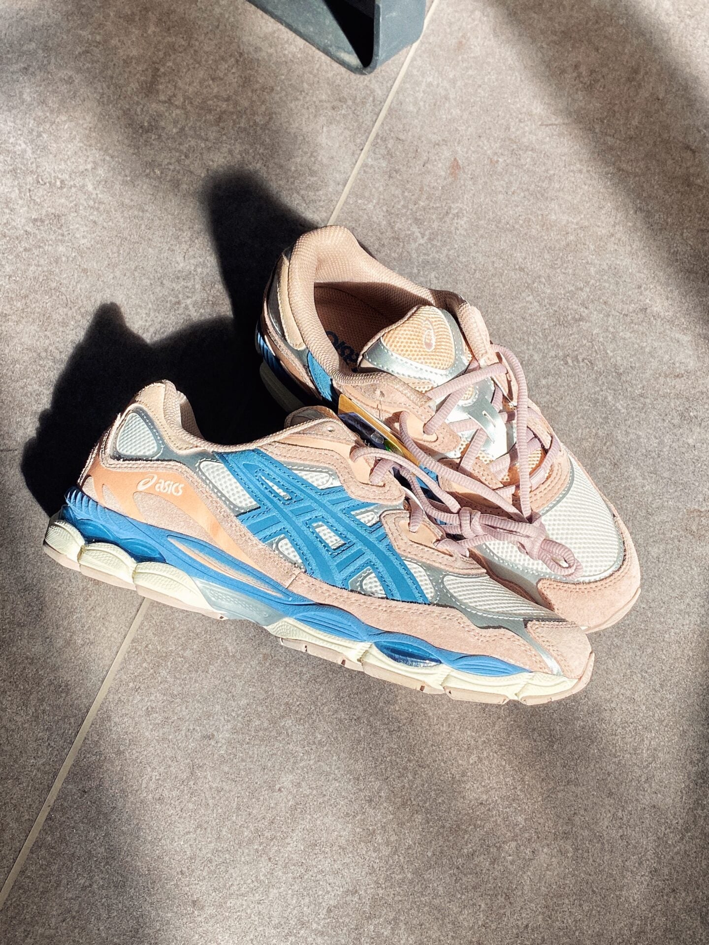 Pair of ASICS Gel-NYC Cream Grey Floss sneakers in cream, grey, and blue tones, styled on a tiled floor with sunlight casting shadows, showcasing exclusive European trainers.