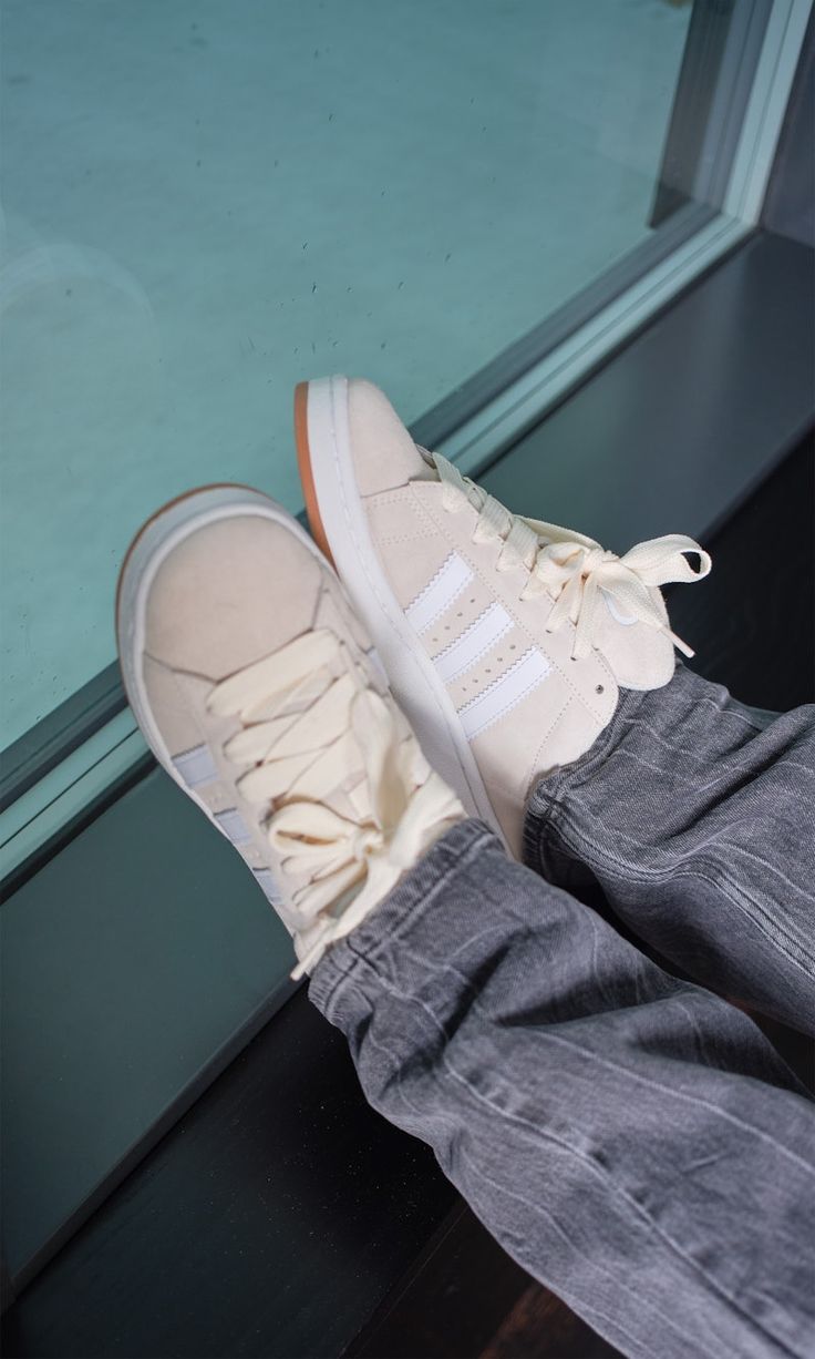 Person wearing Adidas Campus 00s Wonder White sneakers with cream laces and white stripes, paired with grey pants by a window, showcasing exclusive trainers in Europe. The real deal.