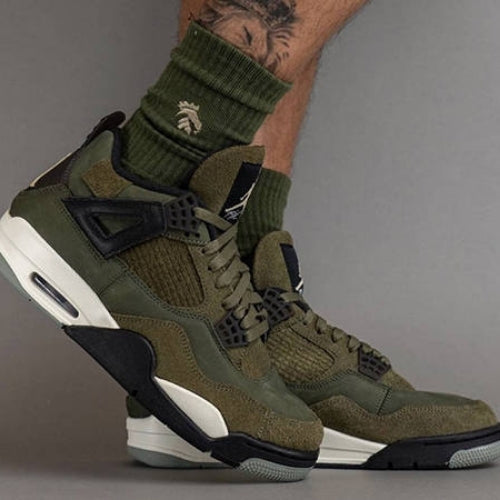 Close-up shot of legs wearing Nike Air Jordan 4 Craft Medium Olive sneakers paired with green ribbed socks, showcasing exclusive sneaker design with mixed textures and muted olive tones.