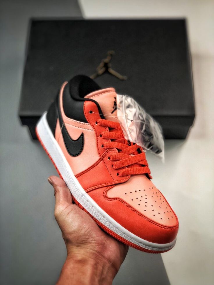 Hand holding an Air Jordan 1 Low Orange Black sneaker featuring orange and black leather uppers, with a white midsole and an orange outsole, against a blurred background with a black box showcasing the iconic Jumpman logo. The real deal exclusive sneakers available in whole Europe.