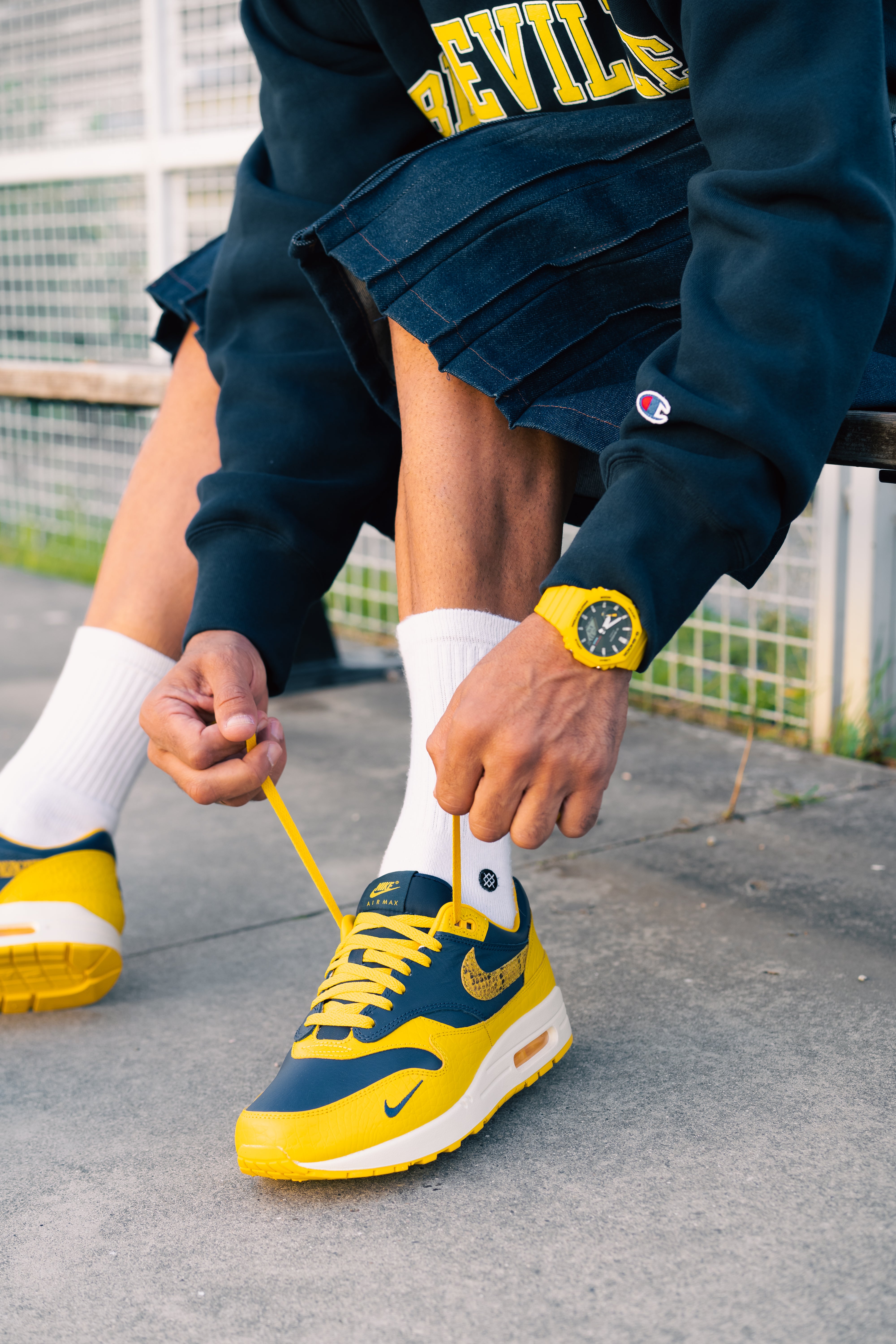 Person tying the laces of a yellow and navy sneaker named Nike Air Max 1 CO.JP Michigan while sitting on a bench, wearing white socks and a dark outfit with a yellow watch, showcasing exclusive sneakers for whole Europe, the real deal.