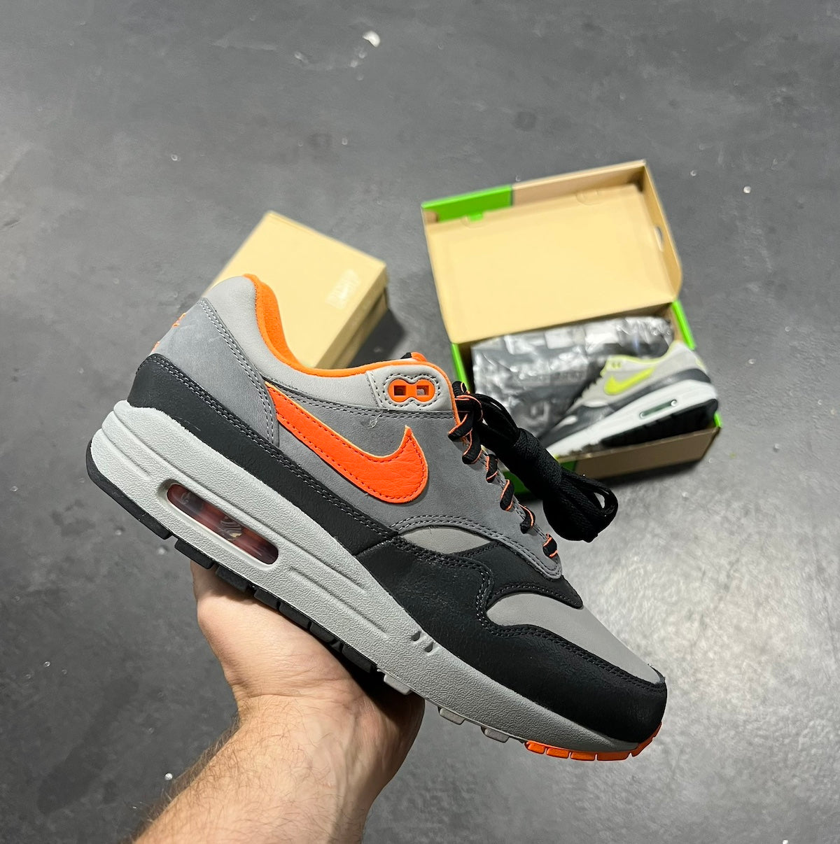 Hand holding a Nike Air Max 1 HUF Brilliant Orange sneaker featuring gray, black, and bright orange accents, with open shoeboxes in the background. The real deal.
