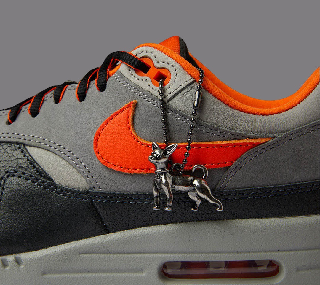 Close-up of a Nike Air Max 1 HUF Brilliant Orange sneaker showcasing its vibrant orange and gray design with a small silver dog charm attached to the laces, representing The real deal exclusive sneakers in Europe.