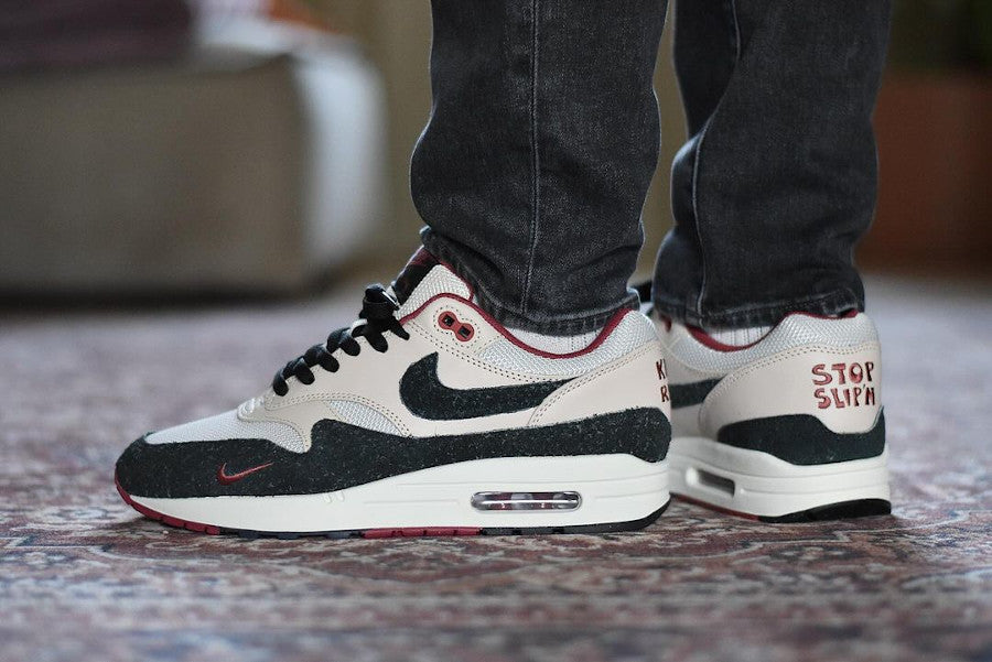 Person wearing Nike Air Max 1 Keep Rippin Stop Slippin 2.0 sneakers in black and white with red accents, standing on a patterned carpet indoors.