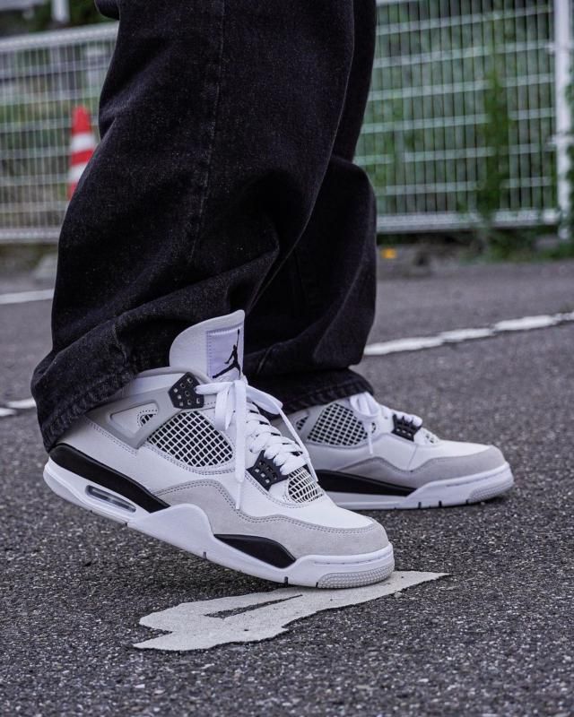 A person wearing Nike Air Jordan 4 Retro Military Black sneakers standing on asphalt, showcasing the detailed design and exclusive style of these sought-after trainers, the real deal.